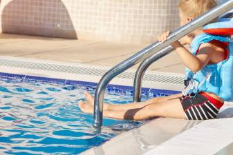 A little boy sits on the side of the pool in a life jacket and is about to dive into the water.