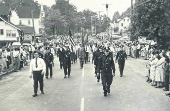 Police Chief John Moeller leads a Memorial Day parade march along Union Valley Road, West Milford, in the 1960s. Frank M. Sell American Legion Post 289 Commander Jack Whitbeck marches next to him. Officers visible behind them include Capt. Jack Ryan, Sgt. Louis Hall and Officers Bill Genader, James Morreale, Bob Deaver and Bill Ohlmeyer. Among Township Council members visible are Township Committeemen Floyd Struble, Warren Brogan and Mayor George Egan. Buildings pictured that no longer are there include the Cracker Barrel Store and Finn family home at left; at right a Flying A Gas Station and the home where seamstress Louise Donnegan had a dry-cleaning business.