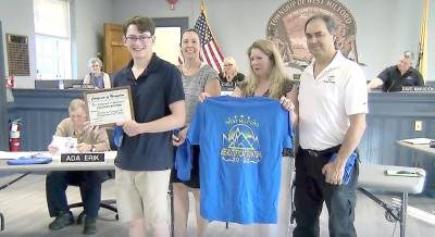 Zachary McPhee, winner of the West Milford Beautification and Recycling Committee shirt design contest, holds his framed certificate. His mother holds a sample blue shirt showing Zachary’s design. The shirt, worn by volunteers in the latest roadside cleanup effort, depicts: five stars for West Milford, a mountain scene and the logo for “Beautification Day 2022.” Also pictured are Zachary’s father and art teacher Jennifer Monego.