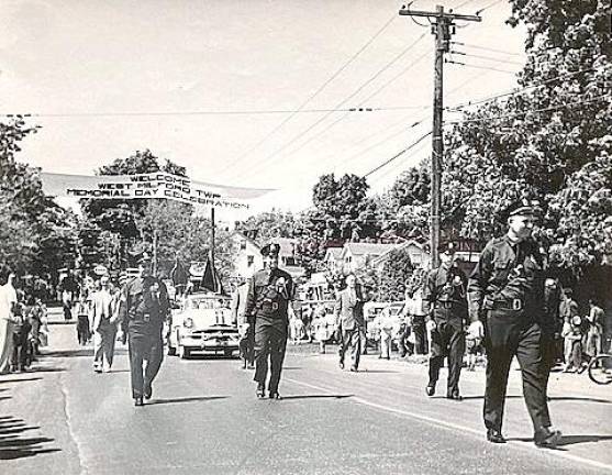 The late Police Chief John Moeller is seen leading the 10th anniversary West Milford Memorial Day Parade in 1954. With Capt. Jack Ryan not in the photo, the remaining members of the five-man full-time police force marching behind the chief are, from left: James Kemble, James Semento and Lou Hall. Township Committee members behind the officers are Bob Little (left) and Chester Pulis (right). Two gas-station signs visible on the right side behind the marchers show Texaco and Esso station signs.
