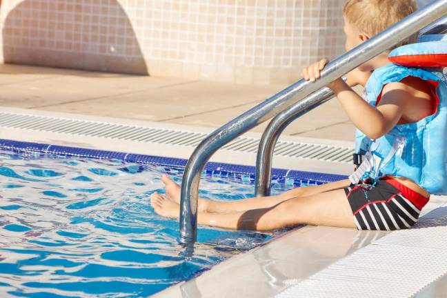 A little boy sits on the side of the pool in a life jacket and is about to dive into the water.