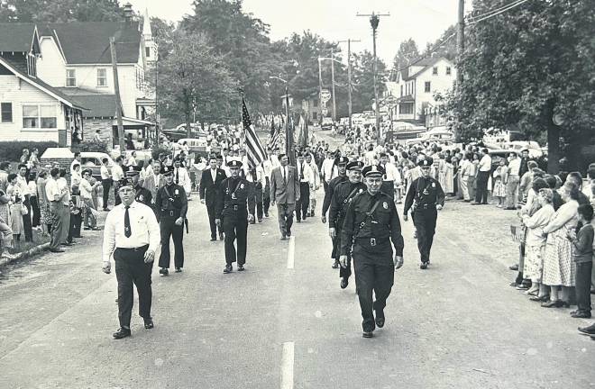 Police Chief John Moeller leads a Memorial Day parade march along Union Valley Road, West Milford, in the 1960s. Frank M. Sell American Legion Post 289 Commander Jack Whitbeck marches next to him. Officers visible behind them include Capt. Jack Ryan, Sgt. Louis Hall and Officers Bill Genader, James Morreale, Bob Deaver and Bill Ohlmeyer. Among Township Council members visible are Township Committeemen Floyd Struble, Warren Brogan and Mayor George Egan. Buildings pictured that no longer are there include the Cracker Barrel Store and Finn family home at left; at right a Flying A Gas Station and the home where seamstress Louise Donnegan had a dry-cleaning business.